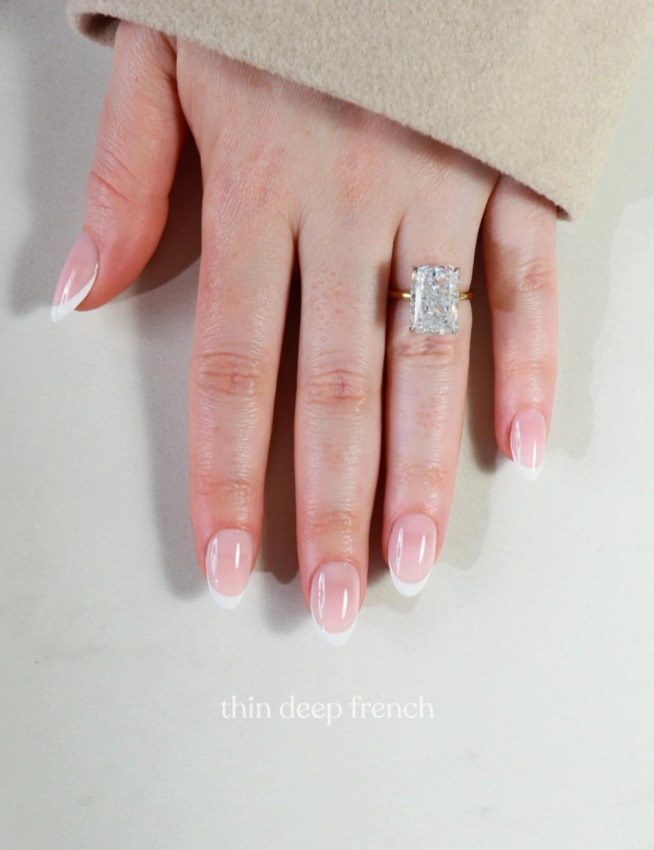 Hand wearing a diamond ring with 'thin deep french' nail design on a neutral background