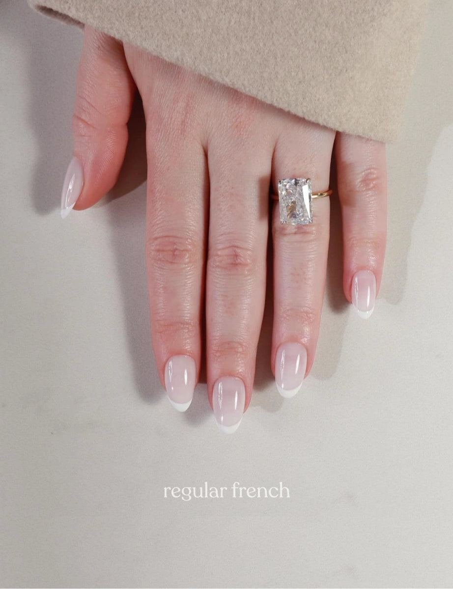 Hand wearing a diamond ring with 'regular french' nail design on a neutral background