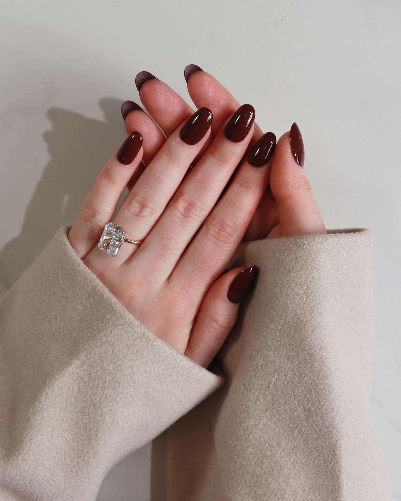 Hand with dark nail polish wearing a ring, wearing a beige sleeve against a neutral background