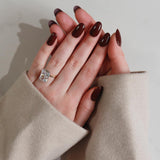 Hand with dark nail polish wearing a ring, wearing a beige sleeve against a neutral background