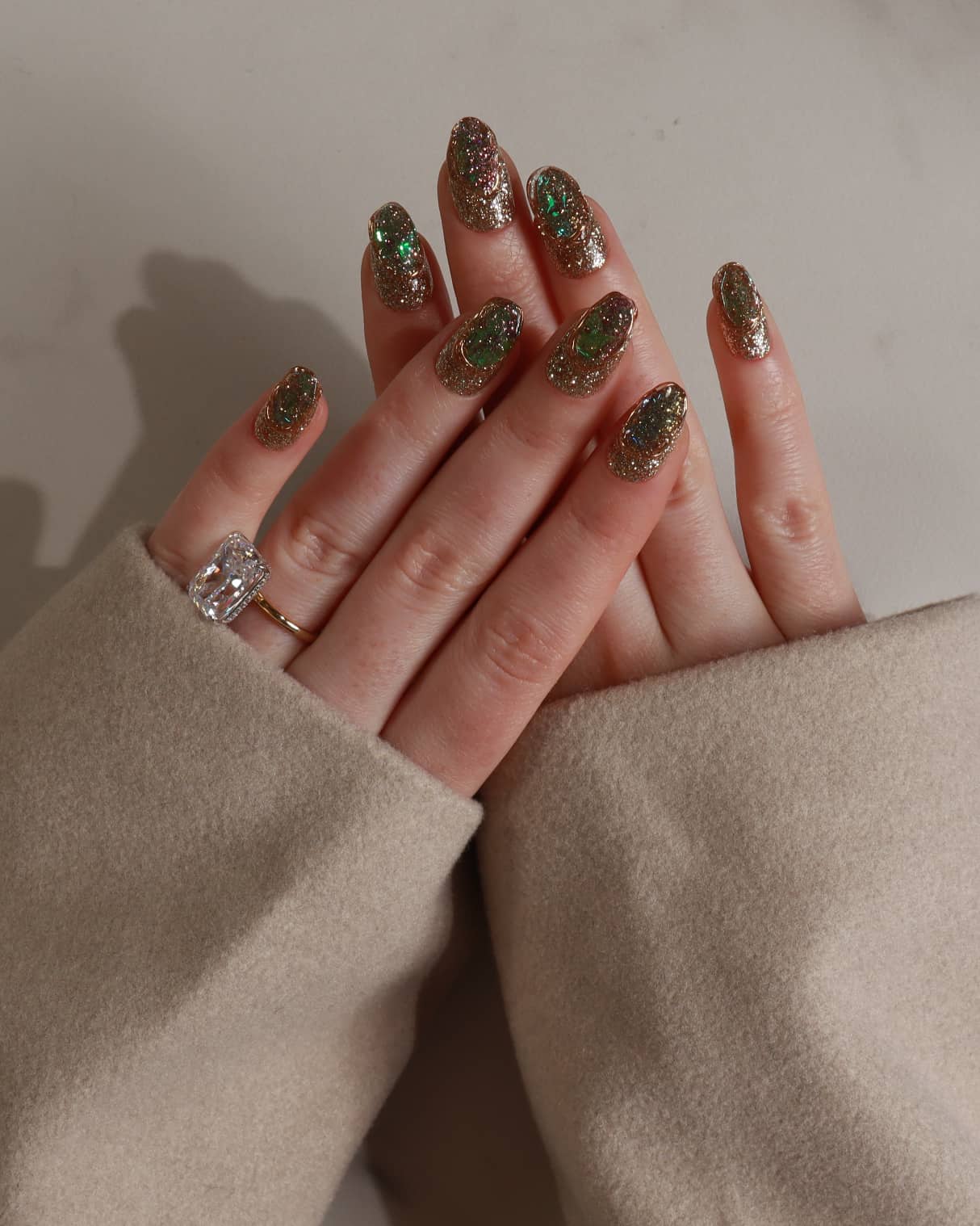 Close-up of a hand with glittery polish and a ring, wearing a beige sleeve.