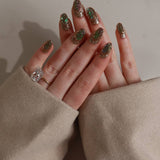 Close-up of a hand with glittery polish and a ring, wearing a beige sleeve.
