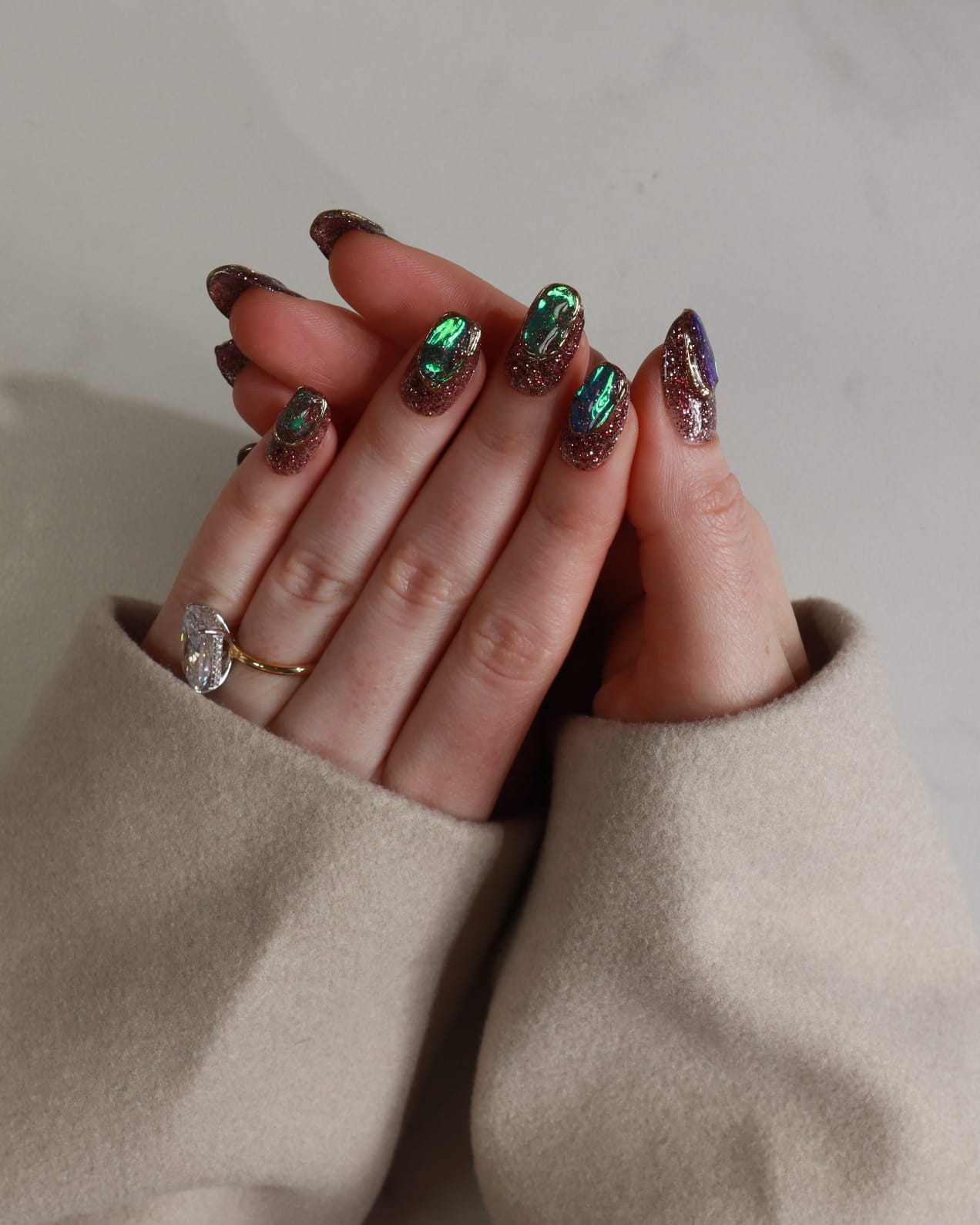 Close-up of a hand with glittery nail polish and rings, wearing a beige sweater.