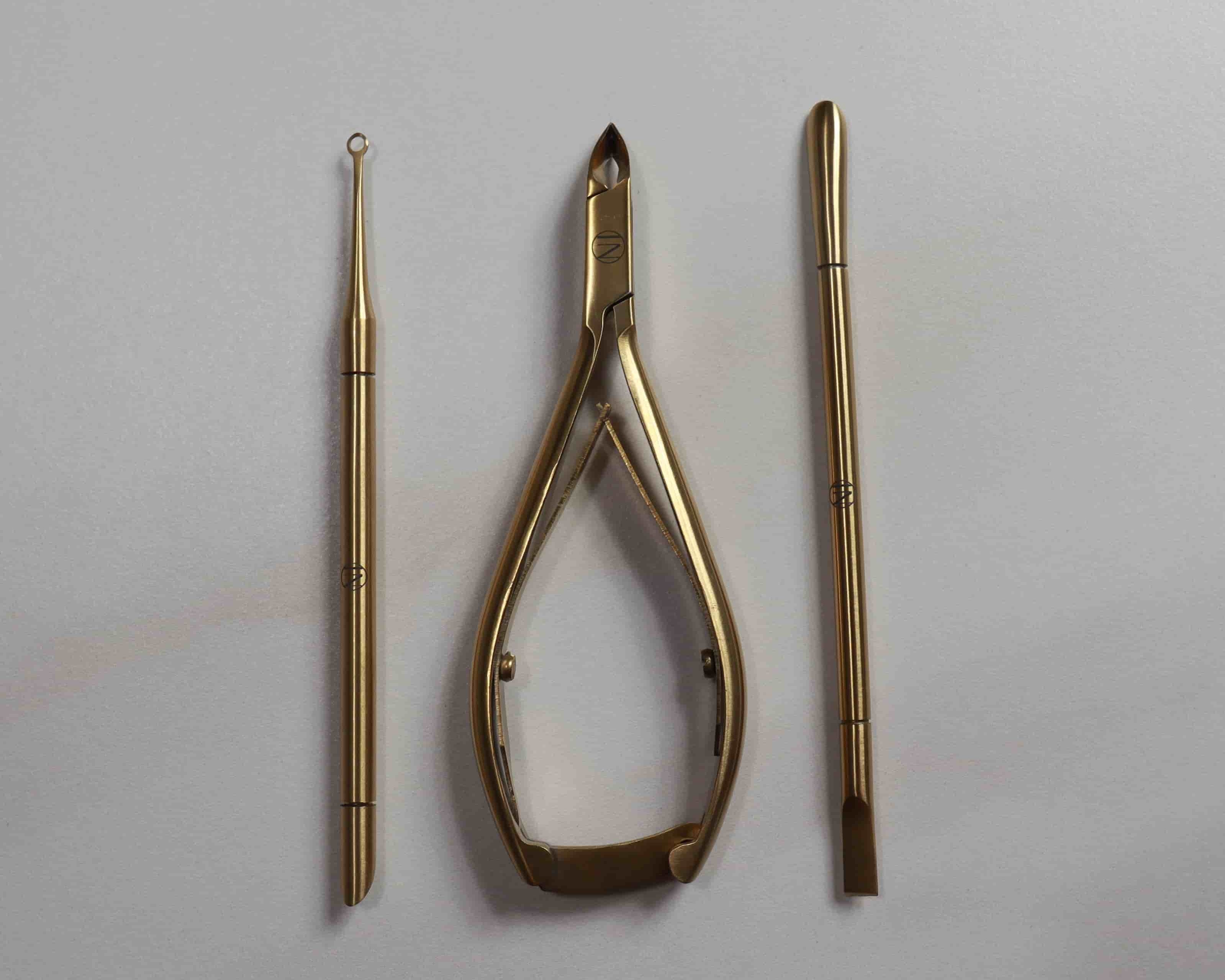Set of gold-colored tools including a pair of cuticle nippers, cuticle pusher, and cuticle loop on a light gray background.