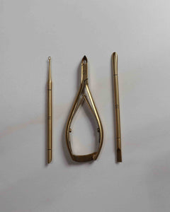 Set of gold-colored tools including a pair of cuticle nippers, cuticle pusher, and cuticle loop on a light gray background.