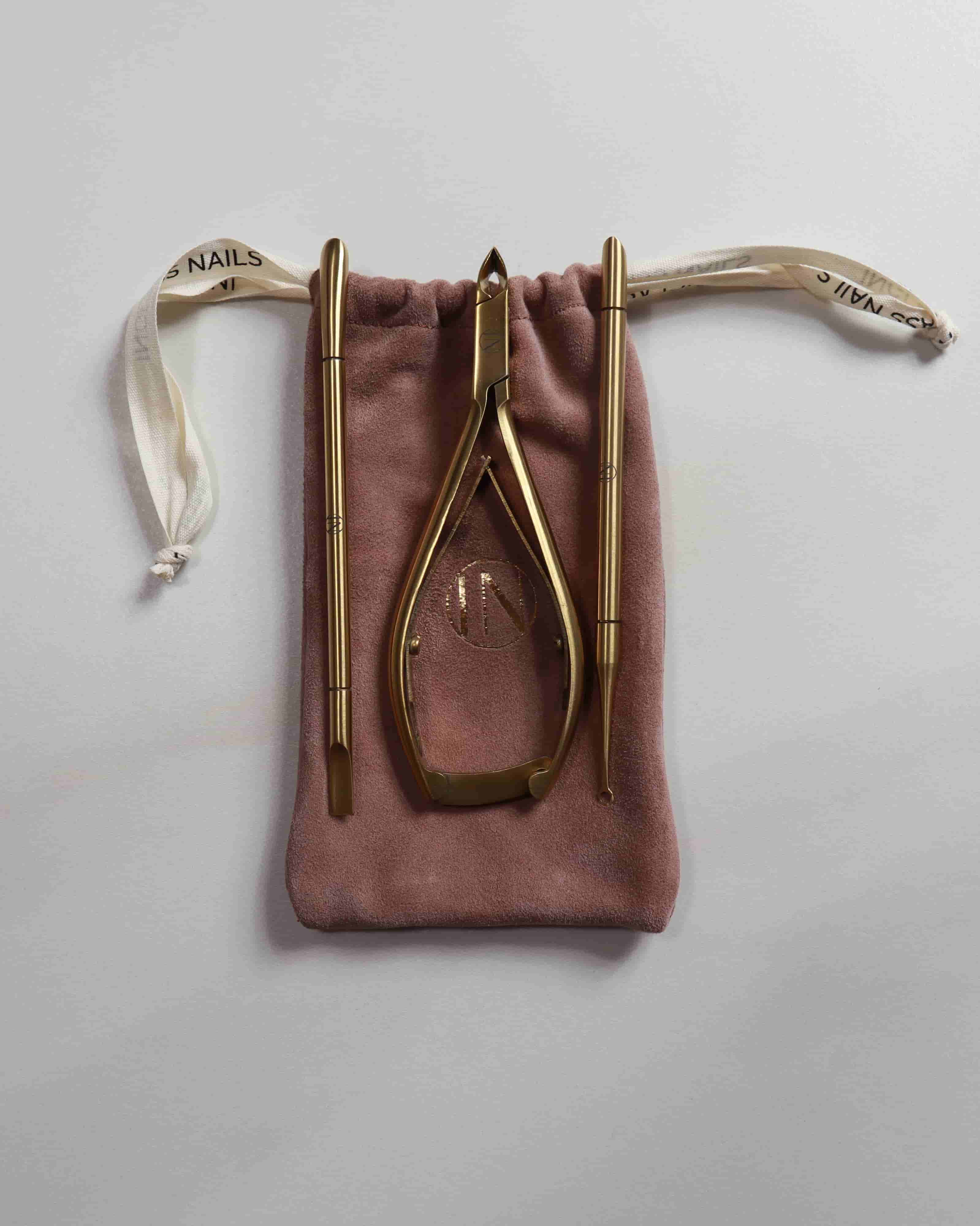 Set of gold nail care tools on a pink drawstring bag against a light gray background