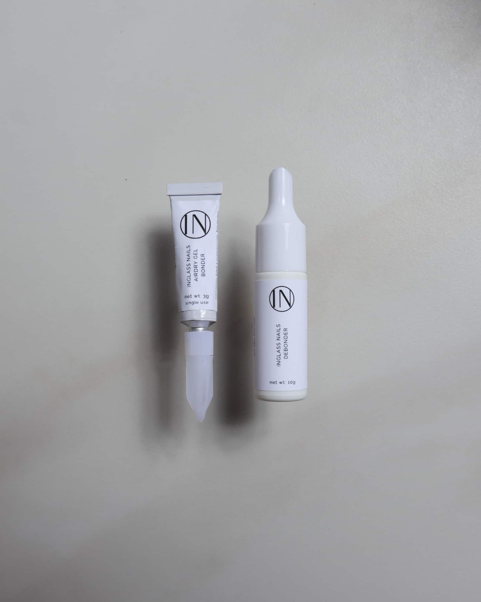Two white tubes from the brand IN, one with a pointed applicator tip and one with a dropper cap, displayed on a neutral light gray background.