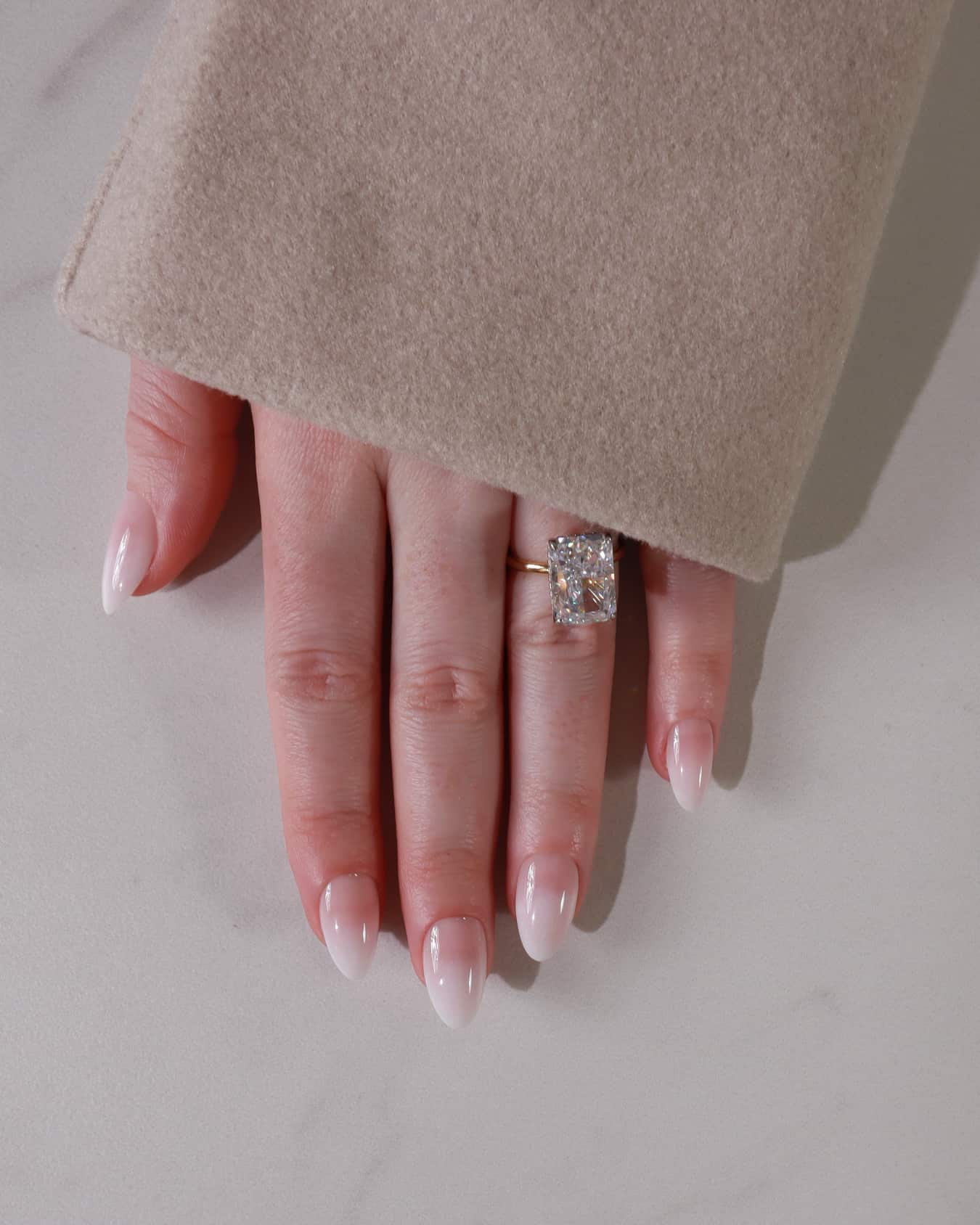 Hand wearing a diamond ring with beige coat sleeve on a neutral background